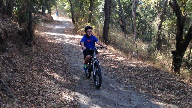 A child riding a bicycle on a dirt path surrounded by trees. The path is partially covered in fallen leaves, and sunlight filters through the foliage. Another cyclist can be seen in the background, and the atmosphere is relaxed and outdoor-oriented. Christmas Hill Garlic Ride mountain bike trail.