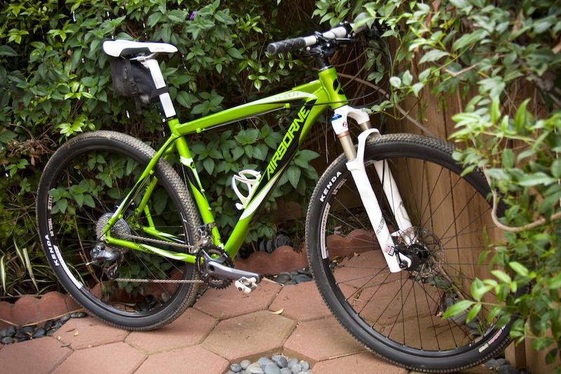 Airborne Goblin: Alt text: A green mountain bike with white accents is parked next to a wooden fence, surrounded by lush green foliage and placed on a brick path.