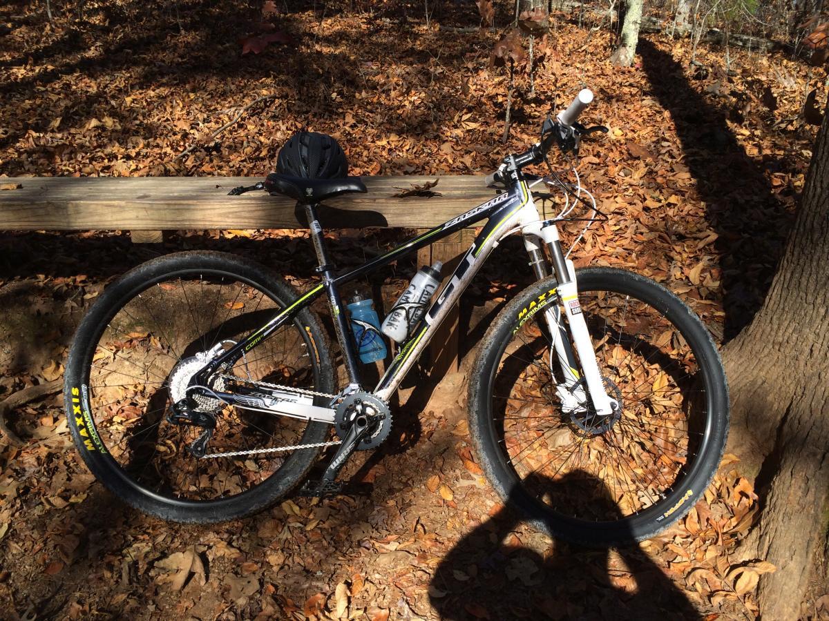 A mountain bike resting on a wooden bench in a forested area, surrounded by fallen leaves. A black helmet is placed on the bench, and a water bottle is attached to the bike. Sunlight filters through the trees, creating dappled shadows on the ground. Blankets Creek mountain bike trail.