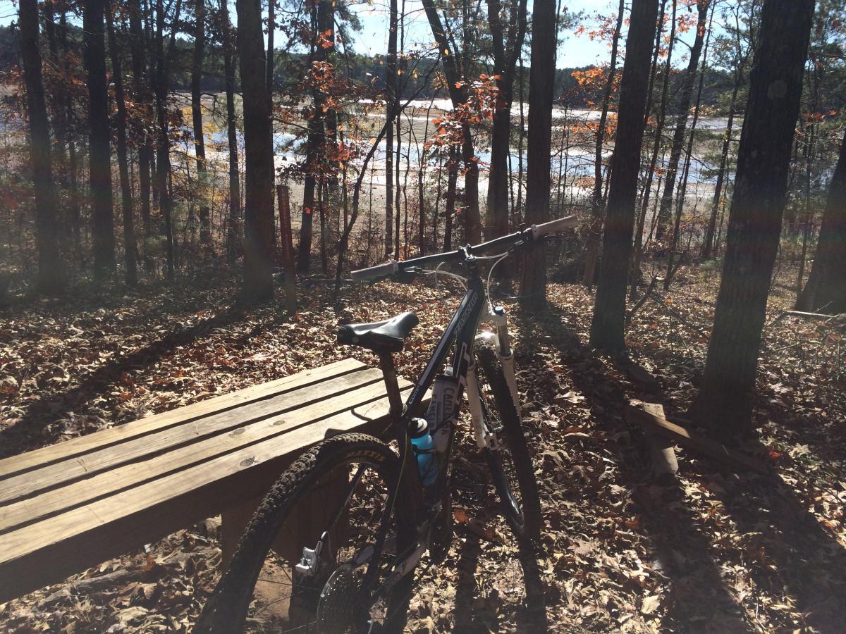 A mountain bike rests on a wooden bench in a sunny forest setting, surrounded by trees with autumn leaves. In the background, a body of water is visible through the trees, suggesting a peaceful outdoor location. Blankets Creek mountain bike trail.