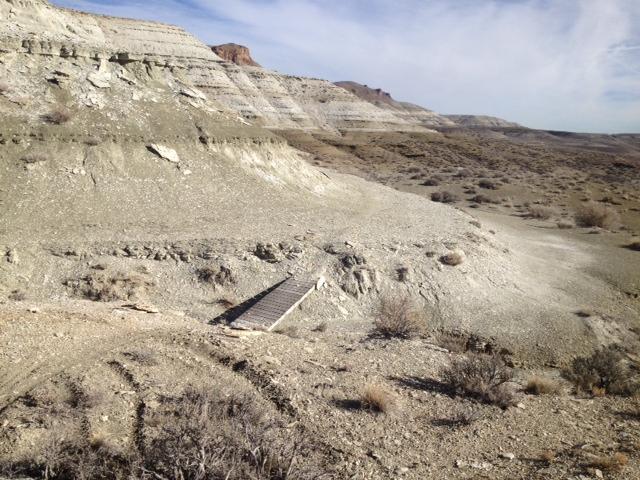 A barren landscape featuring rugged hills and steep slopes, characterized by light-colored rock formations and sparse vegetation. A wooden bridge crosses a shallow ravine in the foreground, leading into the arid terrain under a clear blue sky. Better than Dead mountain bike trail.
