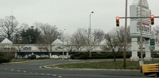 A view of a shopping center on a cloudy day, featuring a row of storefronts with a traffic light in the foreground. The shopping center has distinct tent-like roofs and is surrounded by trees. A roundabout is visible in the foreground, leading to the entrance of the center.