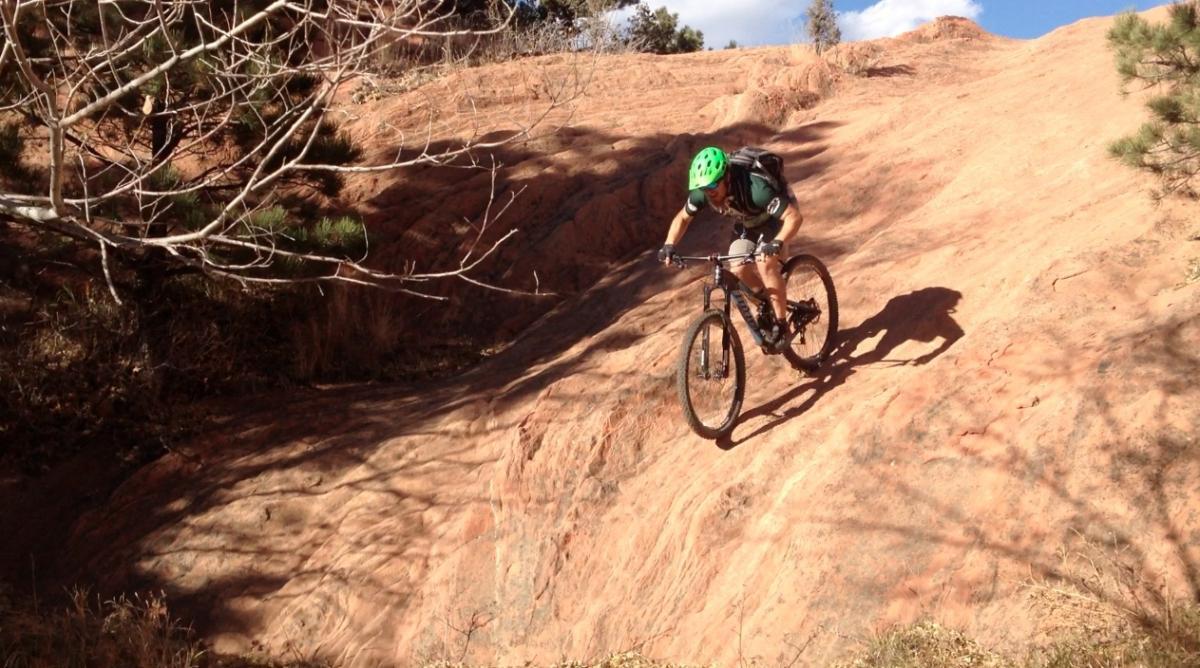 A mountain biker navigating a rocky terrain, wearing a green helmet and a backpack, with trees in the background and a clear blue sky above. Red Rock Canyon mountain bike trail.