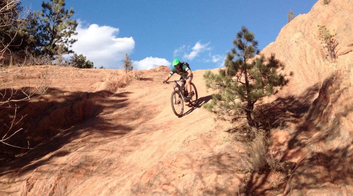 A person riding a mountain bike down a rocky slope, surrounded by trees under a blue sky with scattered clouds. Red Rock Canyon mountain bike trail.