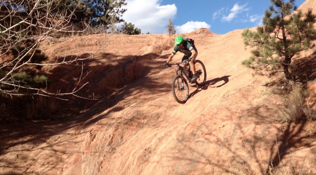 A mountain biker navigates a steep, rocky trail with reddish terrain, surrounded by sparse trees and blue skies. The rider wears a green helmet and is focused on the descent. Red Rock Canyon mountain bike trail.