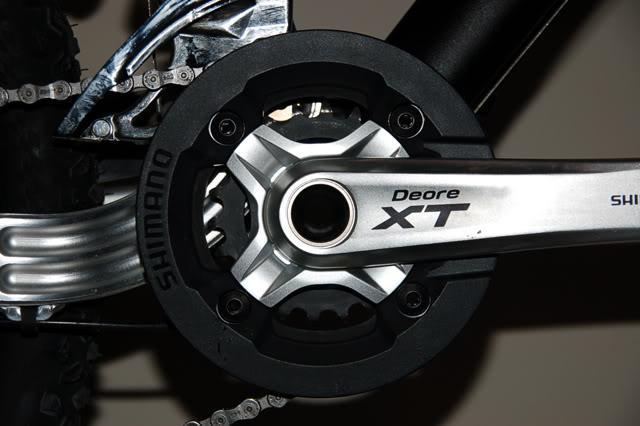 Shimano Saint Bashguard: Close-up of a bicycle's crankset, featuring a Shimano Deore XT component. The metallic crank arm and chainring are prominently displayed, with detailed textures and a black protective cover surrounding the chainring. The background is neutral, emphasizing the bicycle part.