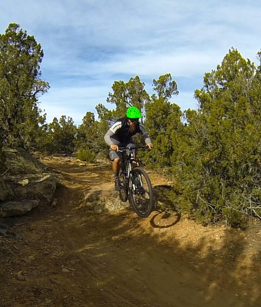 A person wearing a green helmet is riding a mountain bike on a dirt trail surrounded by shrubs and trees. The biker is captured in mid-air, executing a hop over a rocky section of the path, showcasing an active outdoor adventure. The scene is set against a clear sky. Oil Well Flats mountain bike trail.