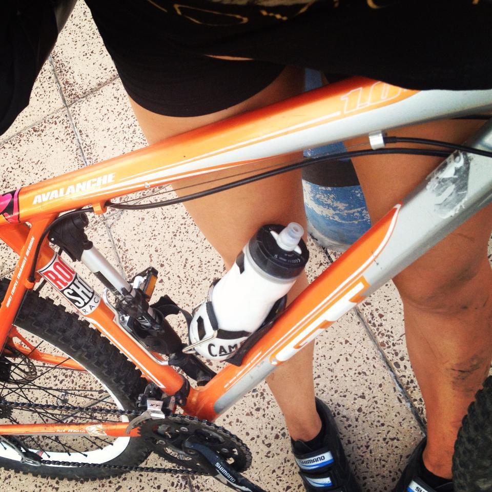 GT Avalanche 1.0 Disc: A close-up view of a mountain bike frame in orange and silver color with a water bottle mounted in the frame. The image shows a person's legs on either side of the bike, wearing black shorts and cycling shoes. The background features a textured surface.
