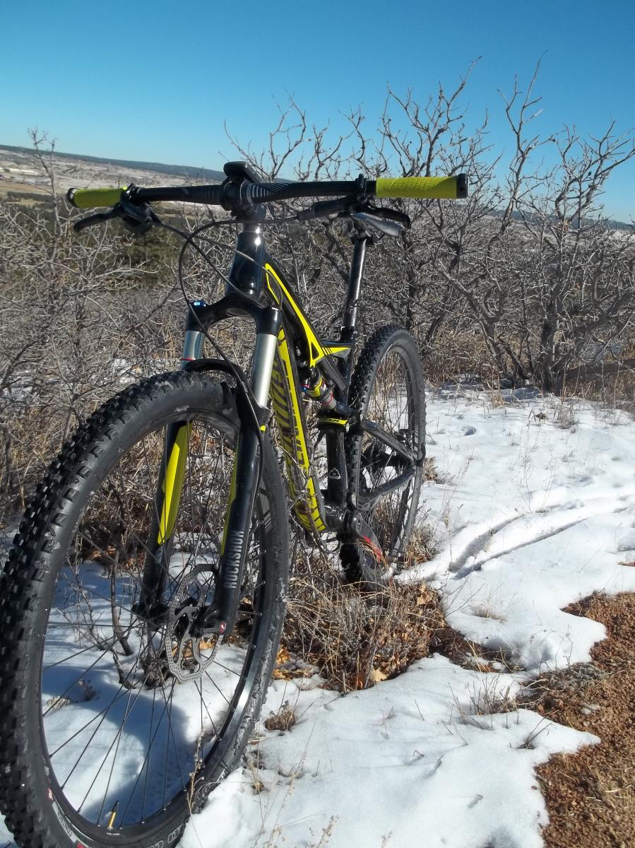 Specialized Camber: A mountain bike with a black and yellow frame parked on a snowy trail surrounded by sparse vegetation and trees. Clear blue sky in the background.