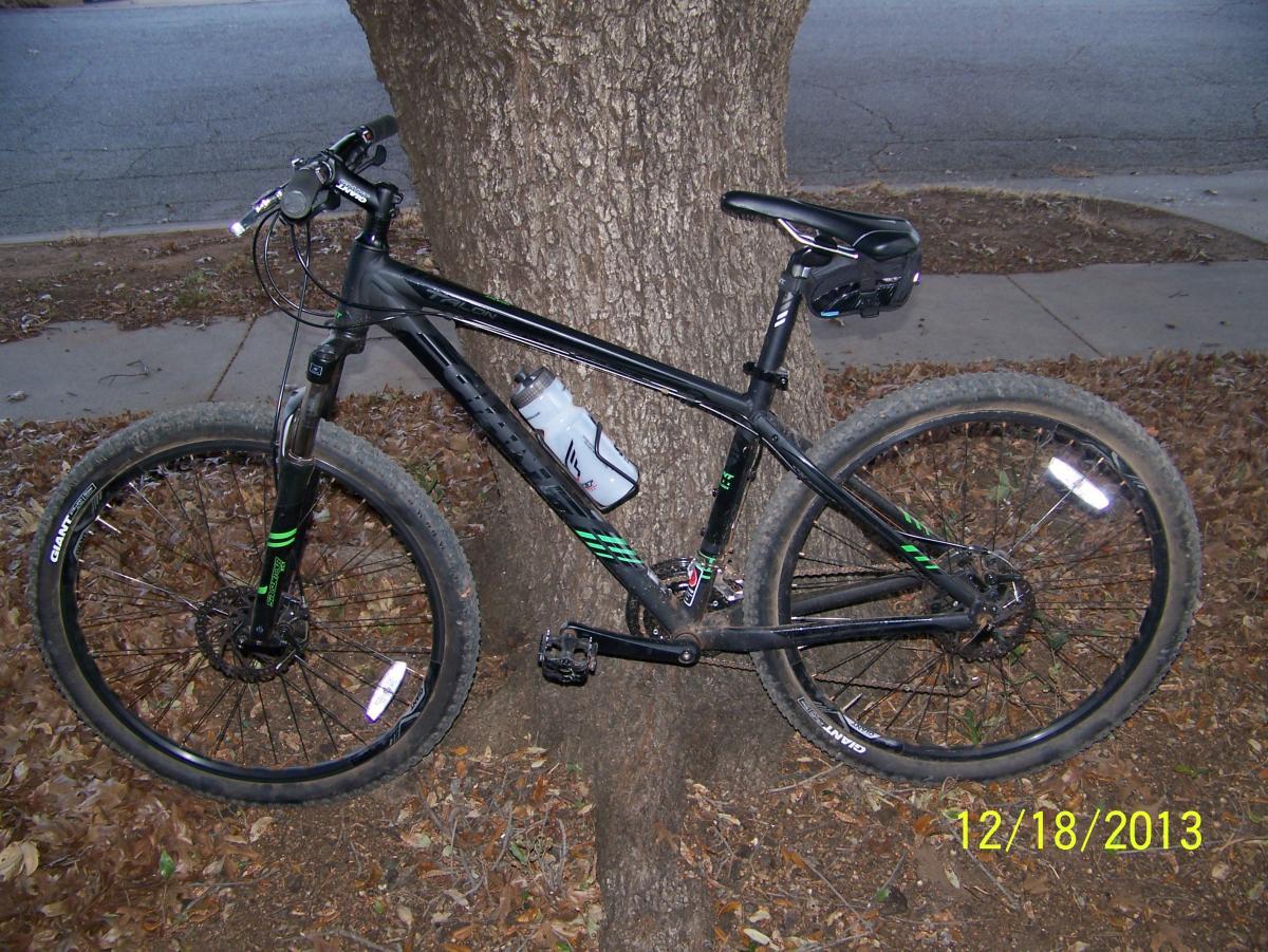 Giant Talon 27.5: A black mountain bike with green accents is leaning against a tree. It has a water bottle attached to the frame and is positioned on a path covered with fallen leaves. The background features a street and a sidewalk, indicating an outdoor setting. The image has a date stamp in the lower right corner showing "12/18/2013."