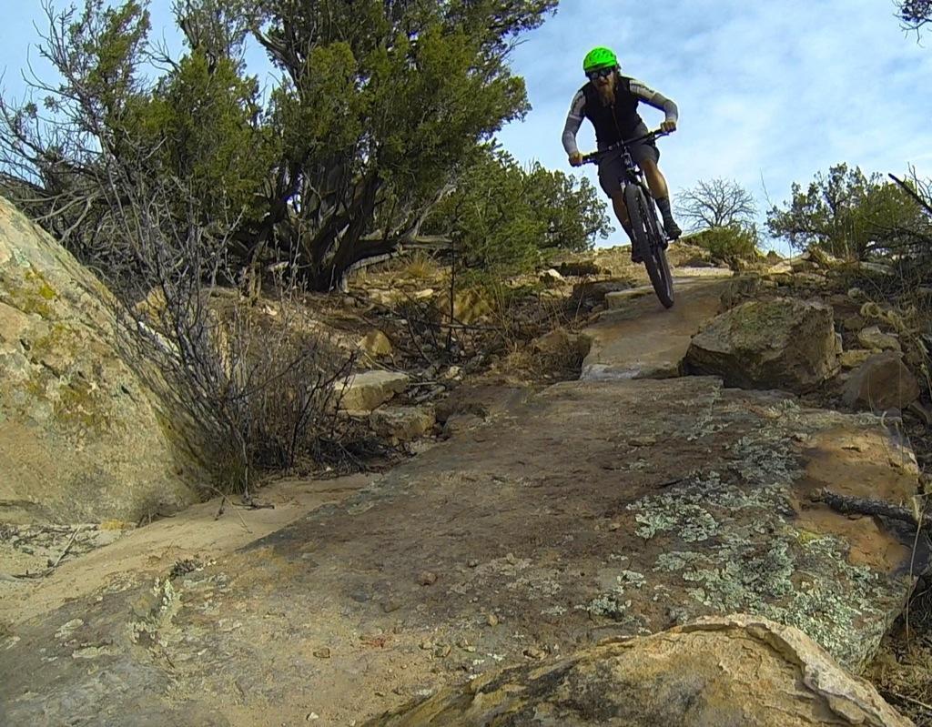 A mountain biker wearing a green helmet and a short-sleeve shirt rides over rocky terrain, navigating a challenging path surrounded by shrubs and trees under a partly cloudy sky. Oil Well Flats mountain bike trail.