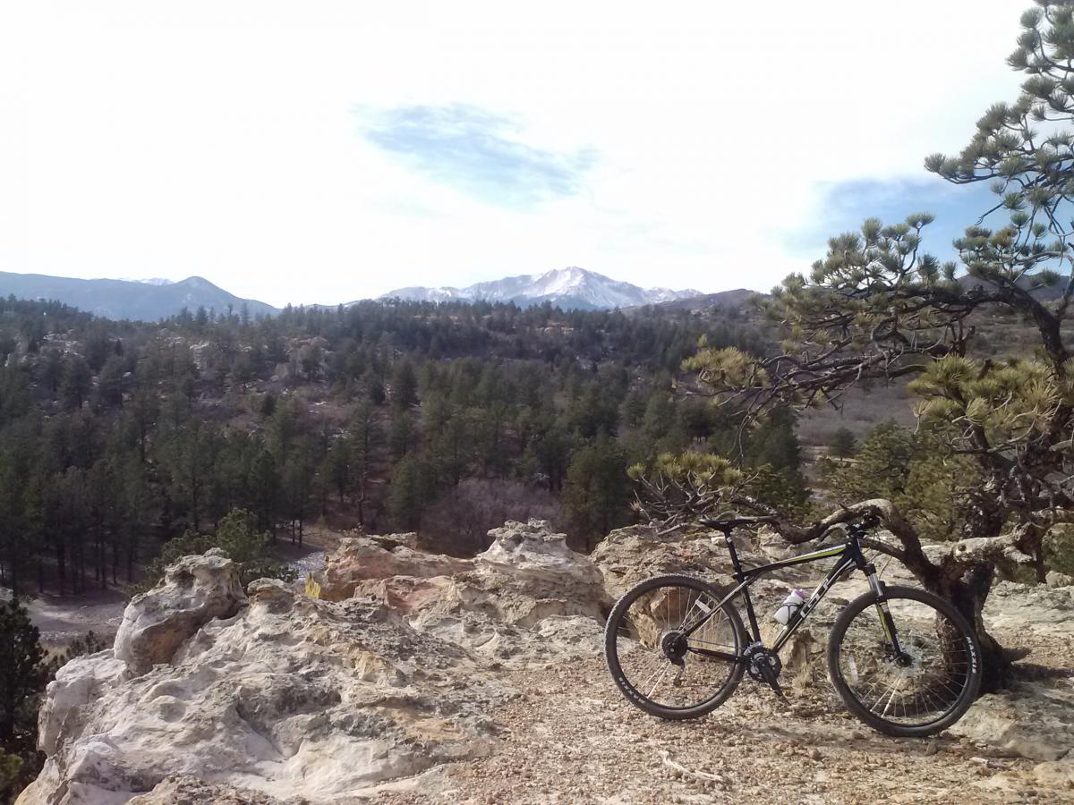 GT Karakoram: A mountain bike is parked on a rocky outcrop, overlooking a vast landscape of evergreen trees and rolling hills, with snow-capped mountains in the distance under a cloudy sky.