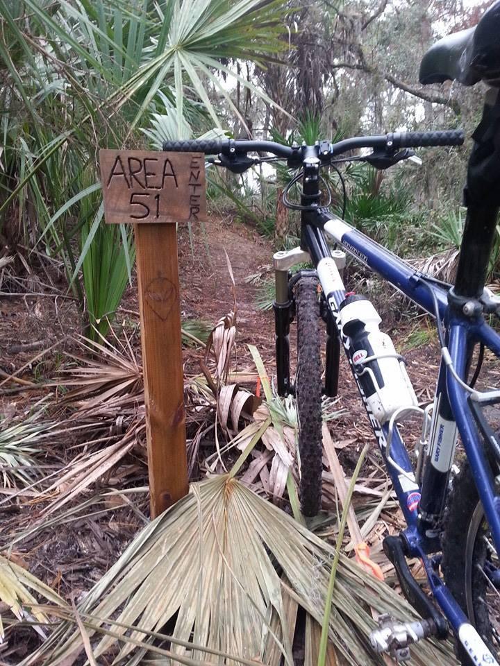 A mountain bike parked beside a wooden sign that reads "AREA 51 ENTER" in a lush, green forest setting. The sign is nestled among palm leaves and other foliage, indicating a pathway into the woods. Carlton Preserve mountain bike trail.