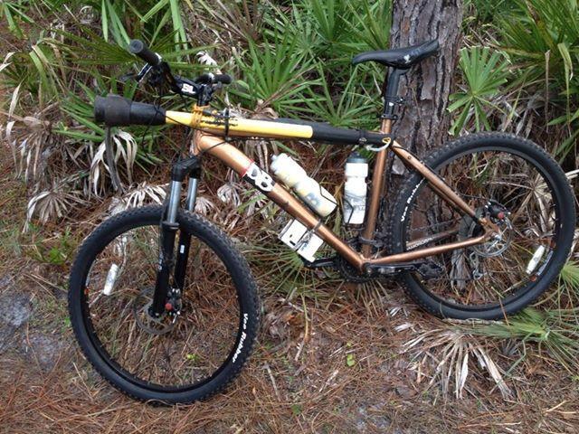 A mountain bike with a brown frame, equipped with two water bottles and other accessories, set against a backdrop of green foliage and tall trees. The bike rests on a dirt path surrounded by natural vegetation. Carlton Preserve mountain bike trail.