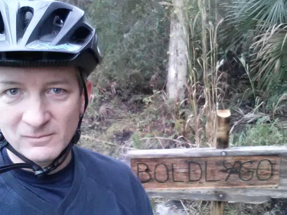 A person wearing a black bicycle helmet stands in front of a wooden sign that reads "BOLDLY GO." The background features dense greenery typical of a wooded area. The individual's expression is focused, and they appear to be ready for an outdoor adventure. Carlton Preserve mountain bike trail.