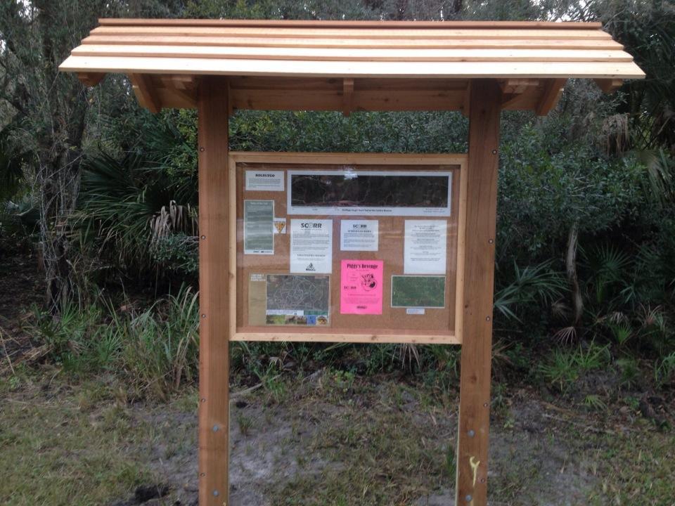 A wooden information board with a slanted roof, displaying various posted materials, including maps, notices, and pamphlets, set in a natural area surrounded by greenery. Carlton Preserve mountain bike trail.