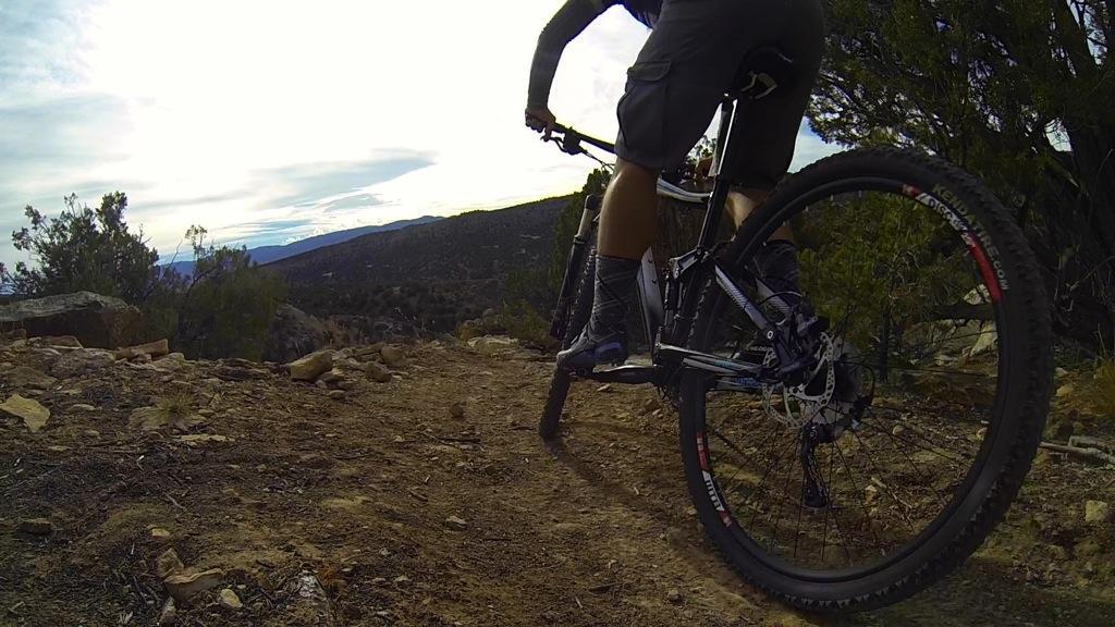 A person riding a mountain bike on a rugged dirt trail, with rocky terrain and sparse vegetation surrounding them. The backdrop features rolling hills under a cloudy sky, creating a scenic outdoor environment. Oil Well Flats mountain bike trail.