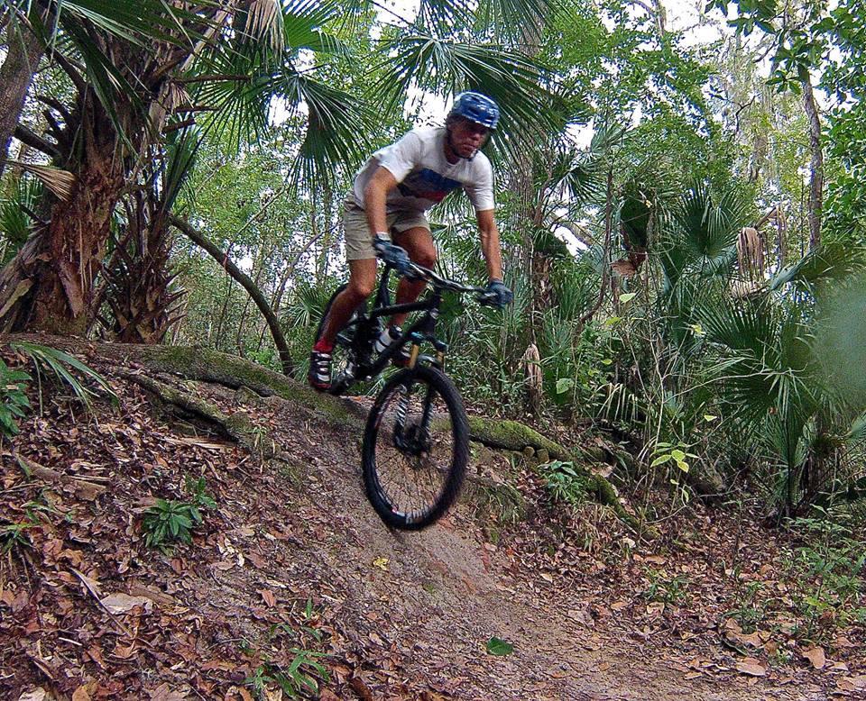 A mountain biker in mid-air performs a jump on a dirt trail surrounded by lush greenery and palm trees in a tropical forest setting. The rider is wearing a helmet and casual riding gear, showcasing an adventurous moment in nature. Chuck Lennon Park mountain bike trail.