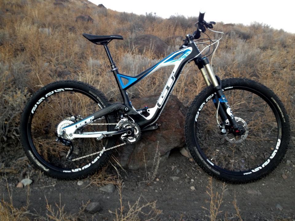 GT Force Carbon Pro: A mountain bike with a blue and silver frame is positioned on a rocky surface, surrounded by dry grass and a rugged terrain. The bike features thick, durable tires and a front suspension fork, indicating its capability for off-road biking.