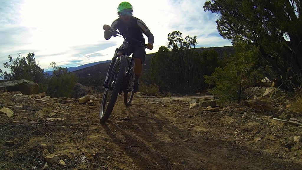 A mountain biker navigating a rocky trail surrounded by trees, with a bright sky in the background. The rider is wearing a green helmet and is captured in motion, highlighting the excitement of off-road biking. Oil Well Flats mountain bike trail.