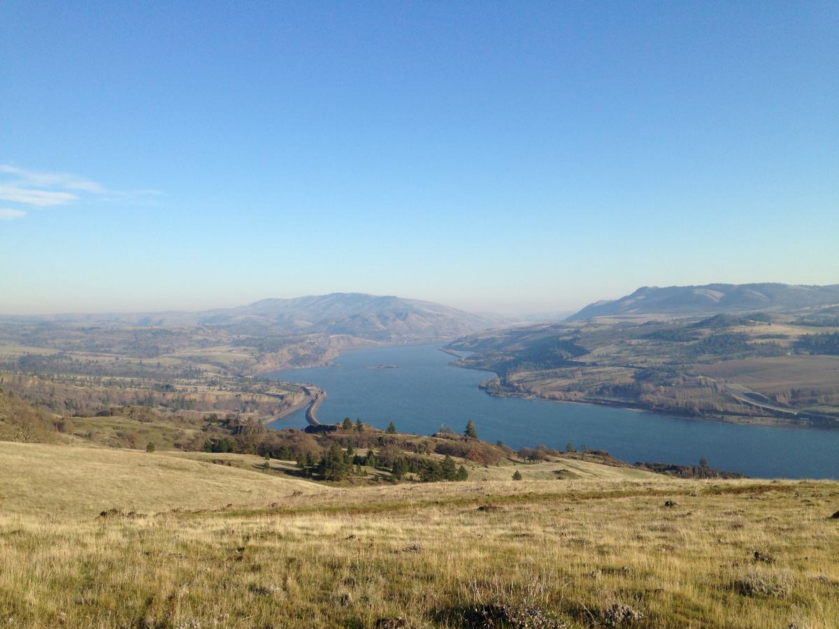 A panoramic view of a river winding through a lush landscape, with hills and mountains in the background under a clear blue sky. The foreground features grassy hills and patches of trees along the riverbanks. Syncline mountain bike trail.