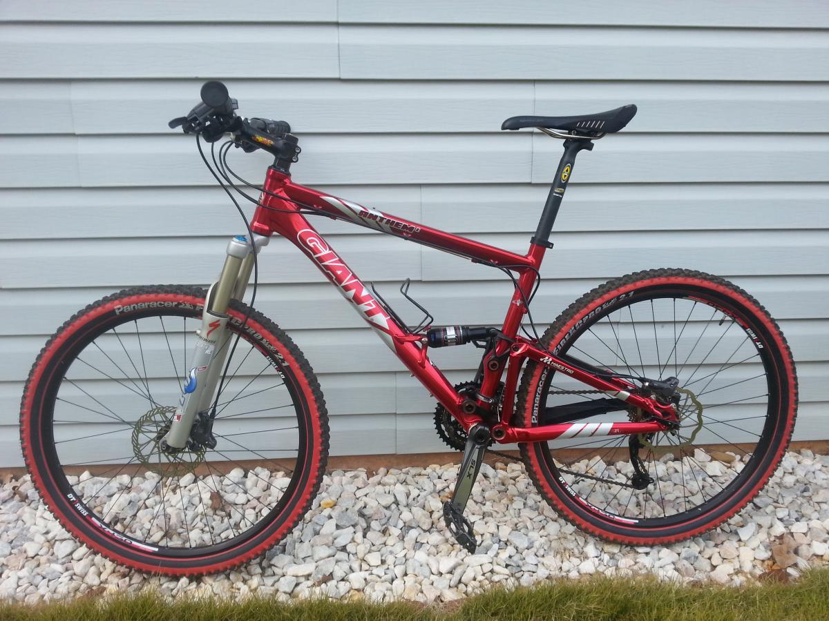 Giant Anthem 2: A red mountain bike with a silver suspension fork, black handlebars, and black tires with red accents, positioned against a light blue wall with gravel and grass in the foreground.
