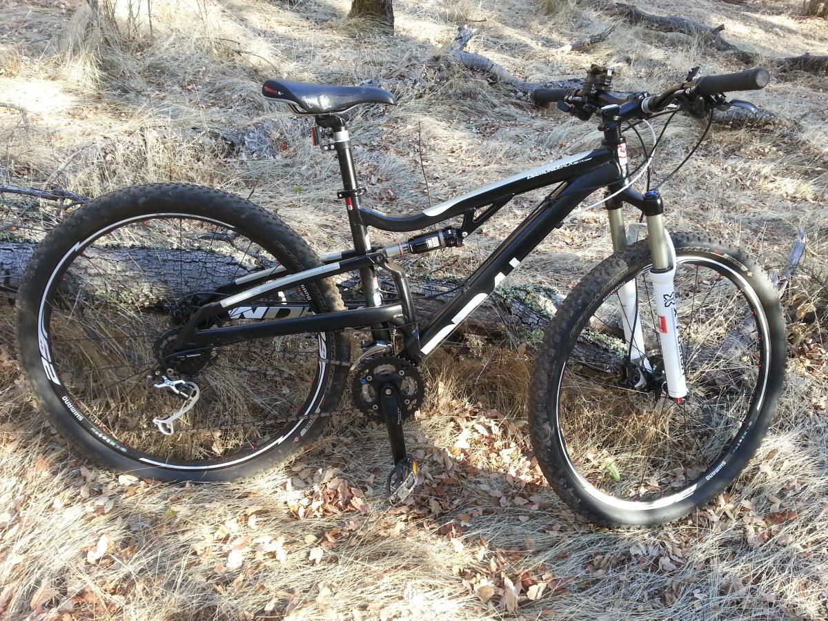 A black mountain bike resting on the ground, surrounded by dry grass and fallen leaves. The bike features thick tires, a suspension fork, and a gear mechanism, with a log visible in the background. Salmon Falls: Sweetwater Loop mountain bike trail.
