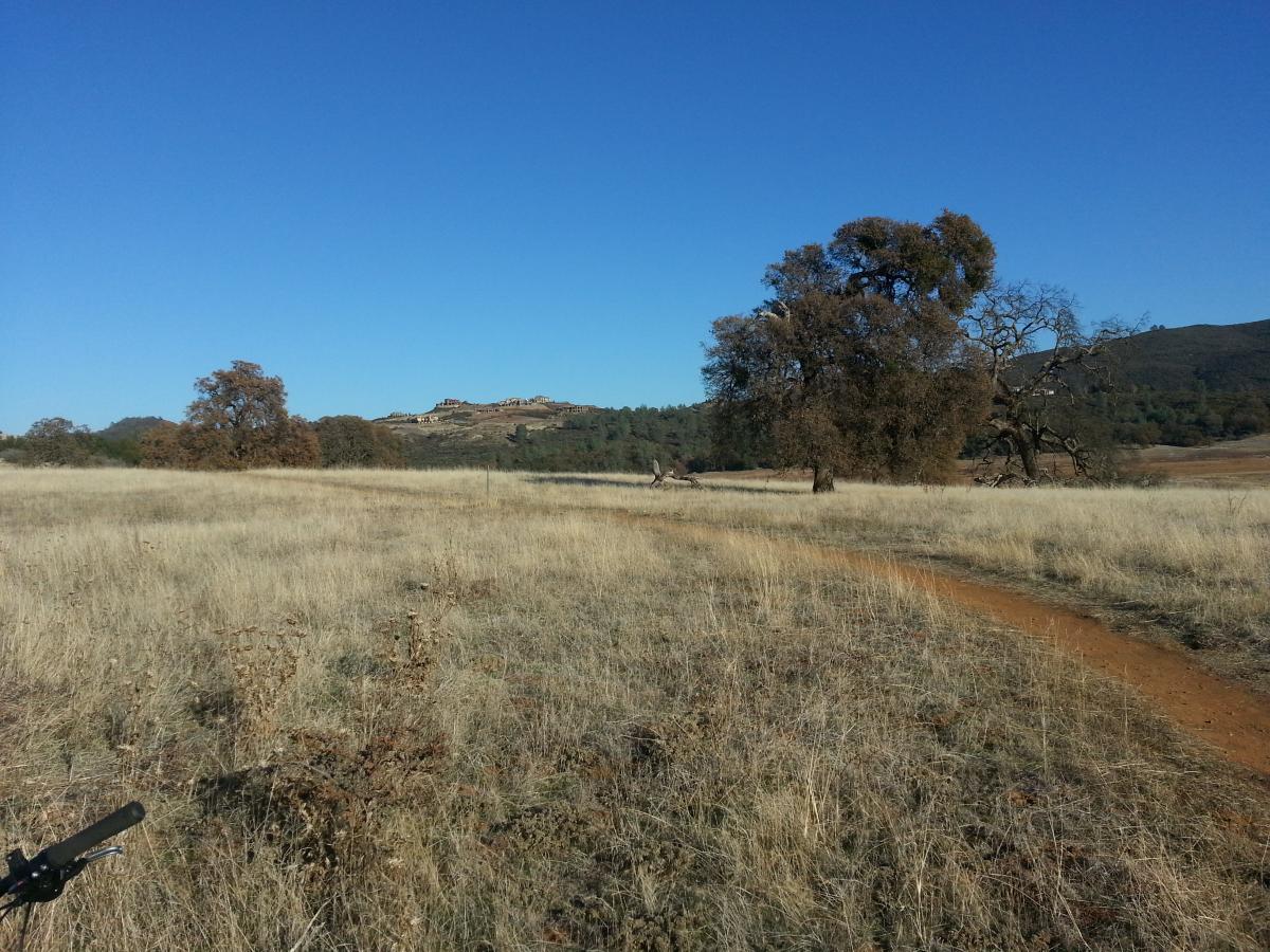 A panoramic view of a dry, grassy field under a clear blue sky, with a dirt path winding through the landscape. A few trees are scattered throughout the scene, and distant hills are visible in the background. Salmon Falls: Sweetwater Loop mountain bike trail.
