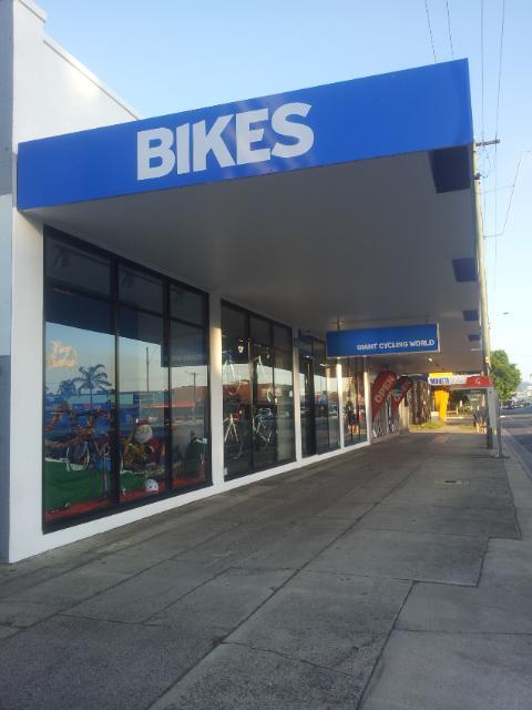 A storefront labeled "BIKES" with large blue signage, featuring large windows displaying various bicycles and cycling accessories. The entrance leads to a store named "Giant Cycling World," located on a sidewalk in a commercial area with clear skies and visible street elements.
