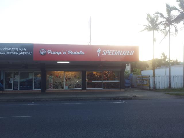Alt text for the image: "Exterior view of a bike shop named 'Pump n' Pedals' with a Specialized sign, featuring large windows showcasing bicycles inside. The storefront is located near a road lined with palm trees."
