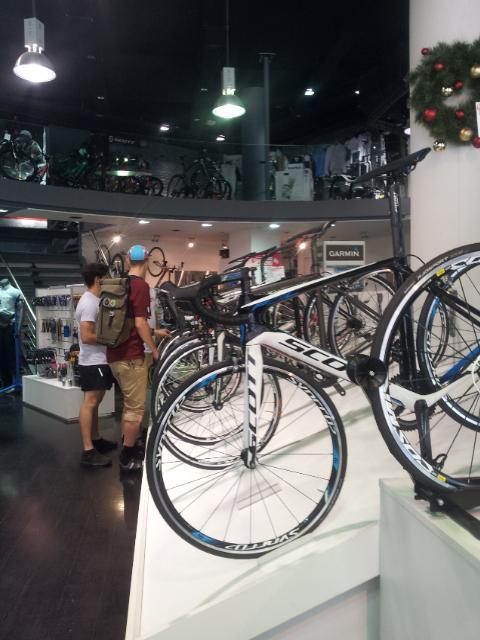 A bike shop interior featuring various bicycles on display, including a close-up of a white and black racing bike in the foreground. Two customers are seen browsing the bicycles, with one wearing a backpack and the other with a baseball cap. The store has a modern design, with stairs leading to an upper level visible in the background.