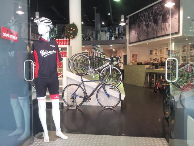 A display window of a bicycle shop featuring a mannequin dressed in cycling gear, including a helmet and jersey. Behind it, several bicycles are showcased, and the interior of the store is visible with a modern design and cycling-themed decor.