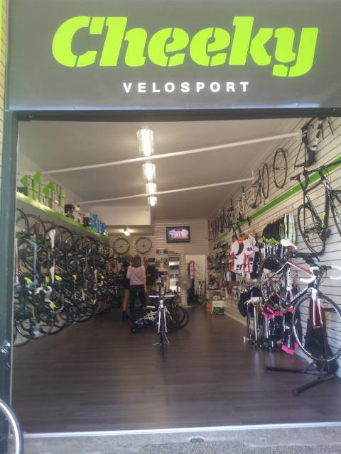Front view of a bicycle shop named "Cheeky Velosport," showcasing a variety of bicycles and cycling accessories on display. The interior has bright lighting, and customers are seen exploring the products. The shop features a sleek design with bikes lined against the walls and a television screen in the background.