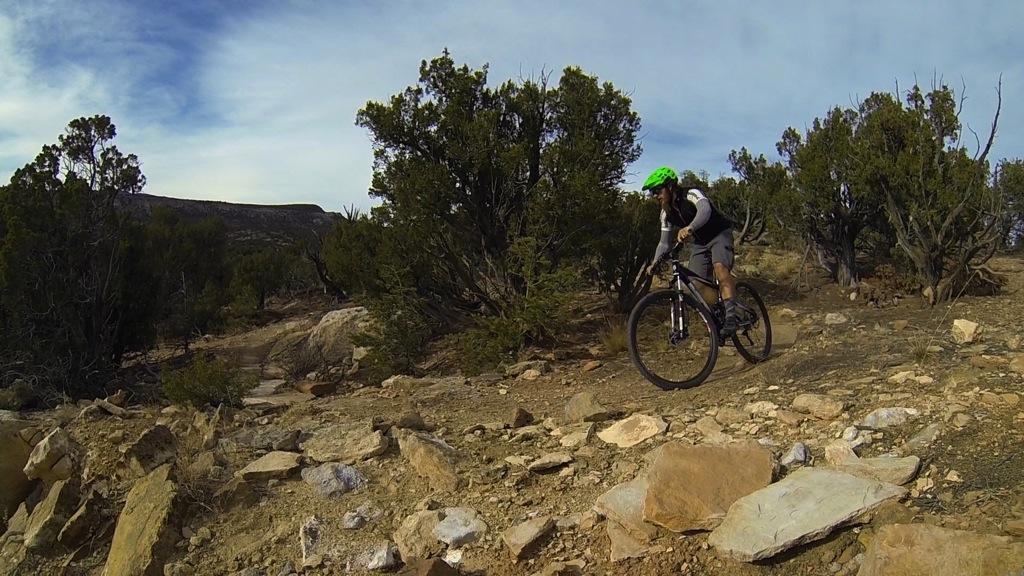 A mountain biker navigating a rocky trail surrounded by shrubs and trees, wearing a green helmet and sports gear, with a mountainous landscape in the background under a partly cloudy sky. Oil Well Flats mountain bike trail.