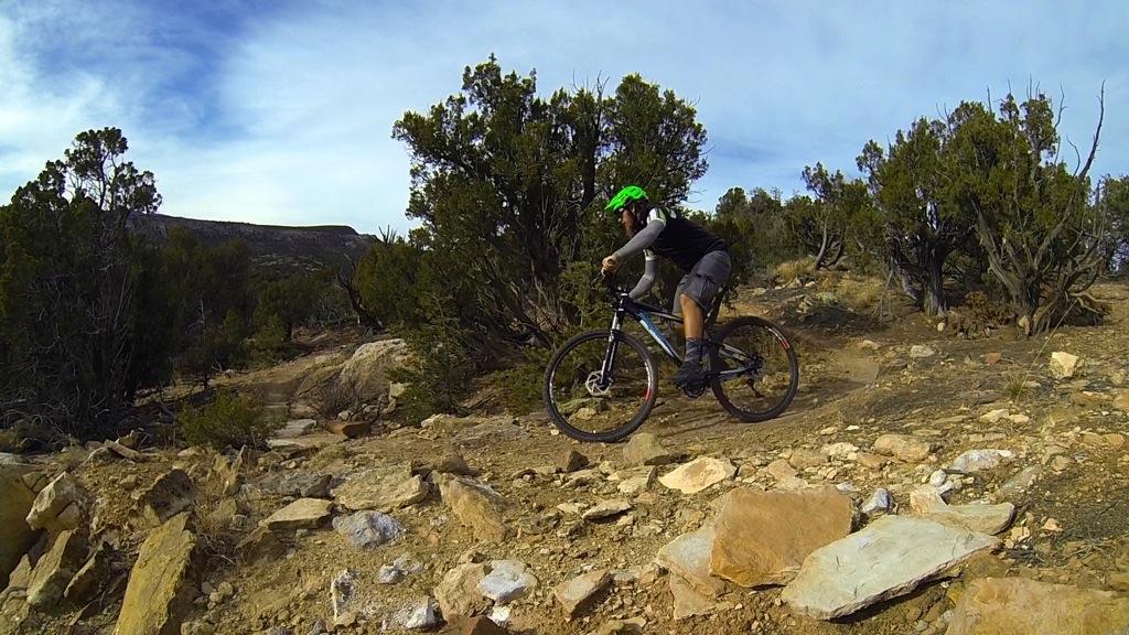 A mountain biker wearing a green helmet navigates a rocky trail surrounded by shrubs and trees. The scene captures the dynamic movement of the rider as they maneuver through the rugged terrain under a cloudy sky. Oil Well Flats mountain bike trail.