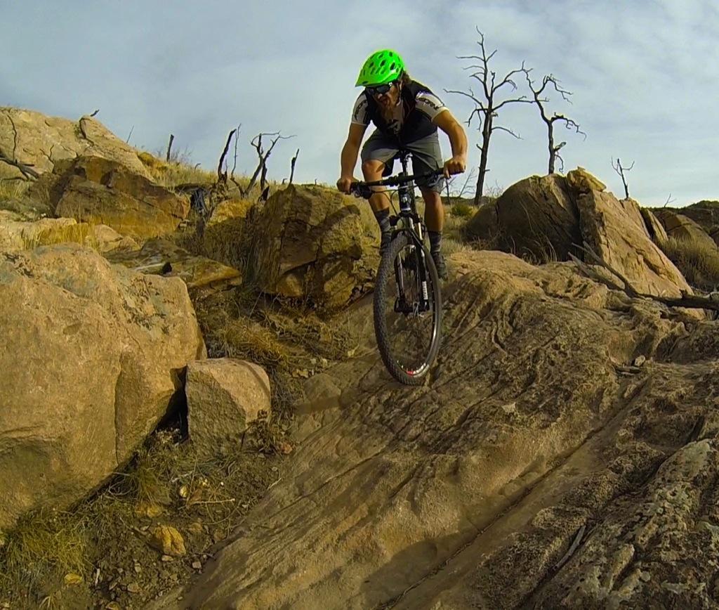 A mountain biker in a bright green helmet navigates a rocky trail, focusing on the path ahead. The rugged terrain features large boulders and dry vegetation, under a partially cloudy sky. Dead trees are visible in the background, suggesting a challenging outdoor environment. Oil Well Flats mountain bike trail.