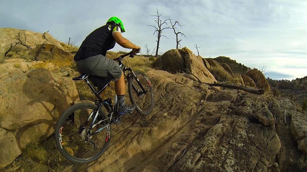 A person wearing a green helmet and black shirt is riding a mountain bike over rocky terrain, navigating steep boulders under a partly cloudy sky. Dead trees are visible in the background, indicating a rugged outdoor environment. Oil Well Flats mountain bike trail.
