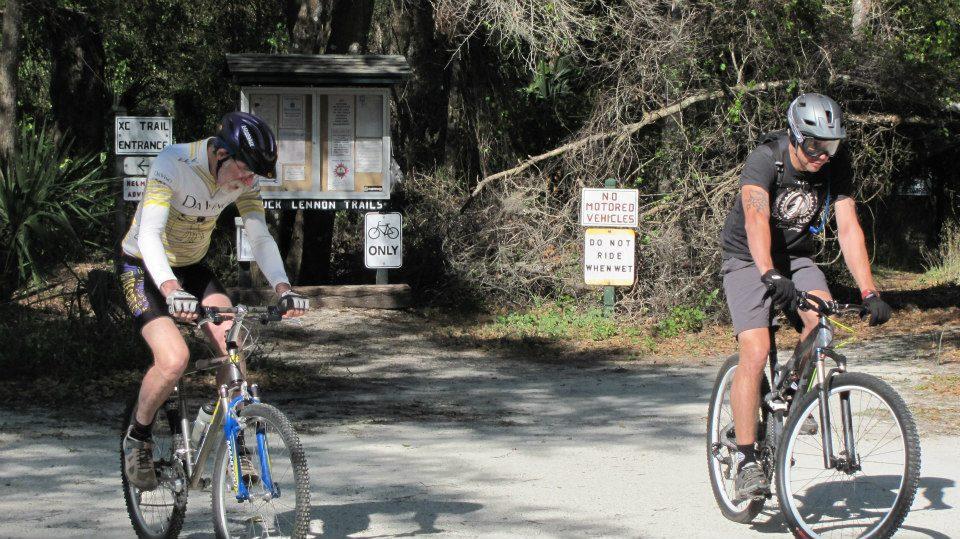 Two mountain bikers are riding on a dirt path near a trail entrance. One cyclist, wearing a yellow cycling jersey, is in the foreground, focused on his ride. The other, dressed in a black shirt and helmet, is slightly behind. In the background, there are signs indicating the start of the trail and rules about no motorized vehicles. Lush greenery surrounds the area, creating a natural setting. Chuck Lennon Park mountain bike trail.