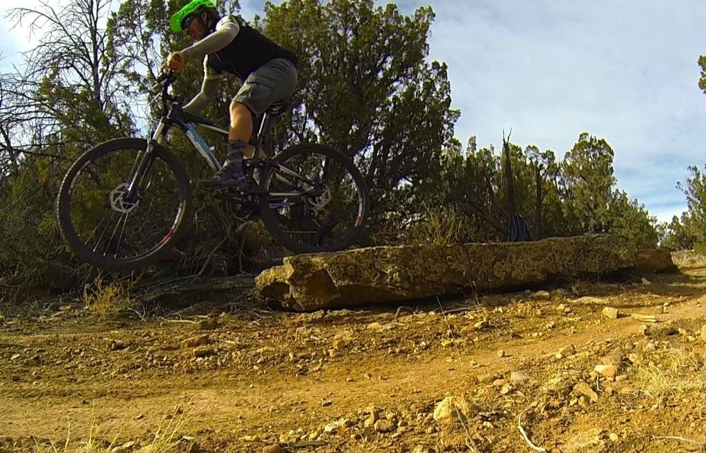 A mountain biker wearing a green helmet and casual gear is airborne as they jump over a large rock on a dirt trail surrounded by trees and shrubs. The scene captures the dynamic action of the jump against a backdrop of natural vegetation under a partly cloudy sky. Oil Well Flats mountain bike trail.
