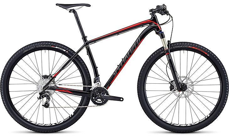 Specialized STUMPJUMPER COMP HT: A modern mountain bike featuring a black frame with red accents, designed for off-road cycling. It has rugged tires, a front suspension fork, and a gear system visible on the frame for variable speeds. The bike is positioned to showcase its sleek design and functionality.