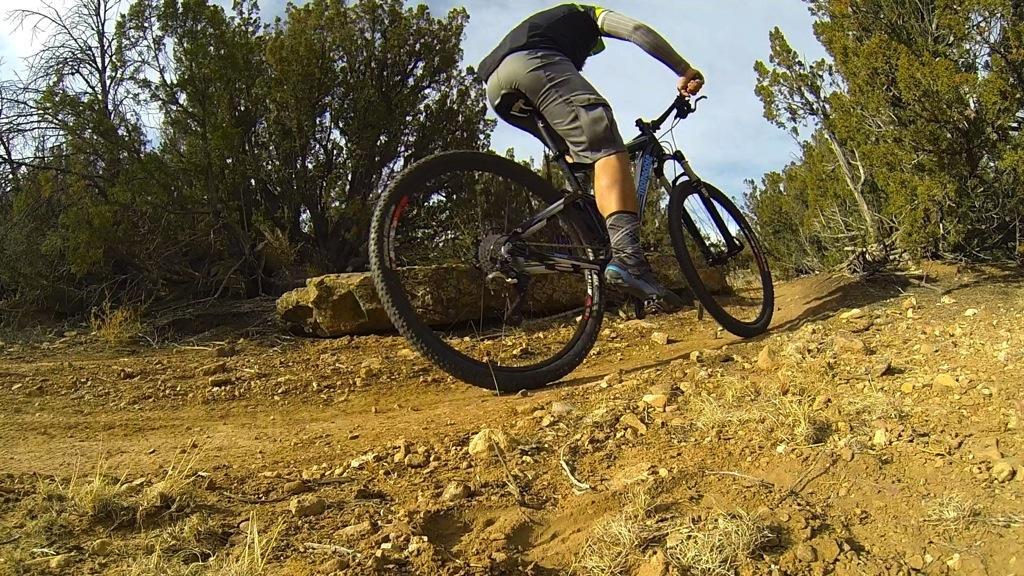 A cyclist riding a mountain bike on a dirt trail surrounded by shrubs and trees, with an emphasis on the bike's rear wheel and the cyclist's lower body. Oil Well Flats mountain bike trail.