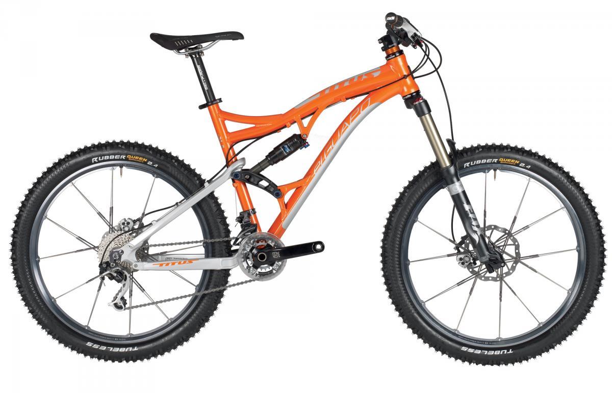 Titus El Guapo: An orange and silver mountain bike with thick tires, a suspension system, and a sleek frame positioned against a plain background. The bike features disc brakes and a variety of gears, making it suitable for off-road cycling.