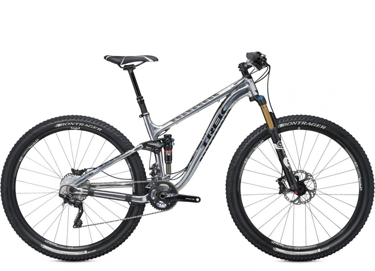Trek Fuel EX 9 29: A silver mountain bike with a sleek design, featuring wide tires, front suspension, and detailed componentry suitable for off-road cycling. The bike is presented at an angle, showcasing its frame, gears, and suspension system. The handlebars show a digital display and brake setup, emphasizing its advanced features.