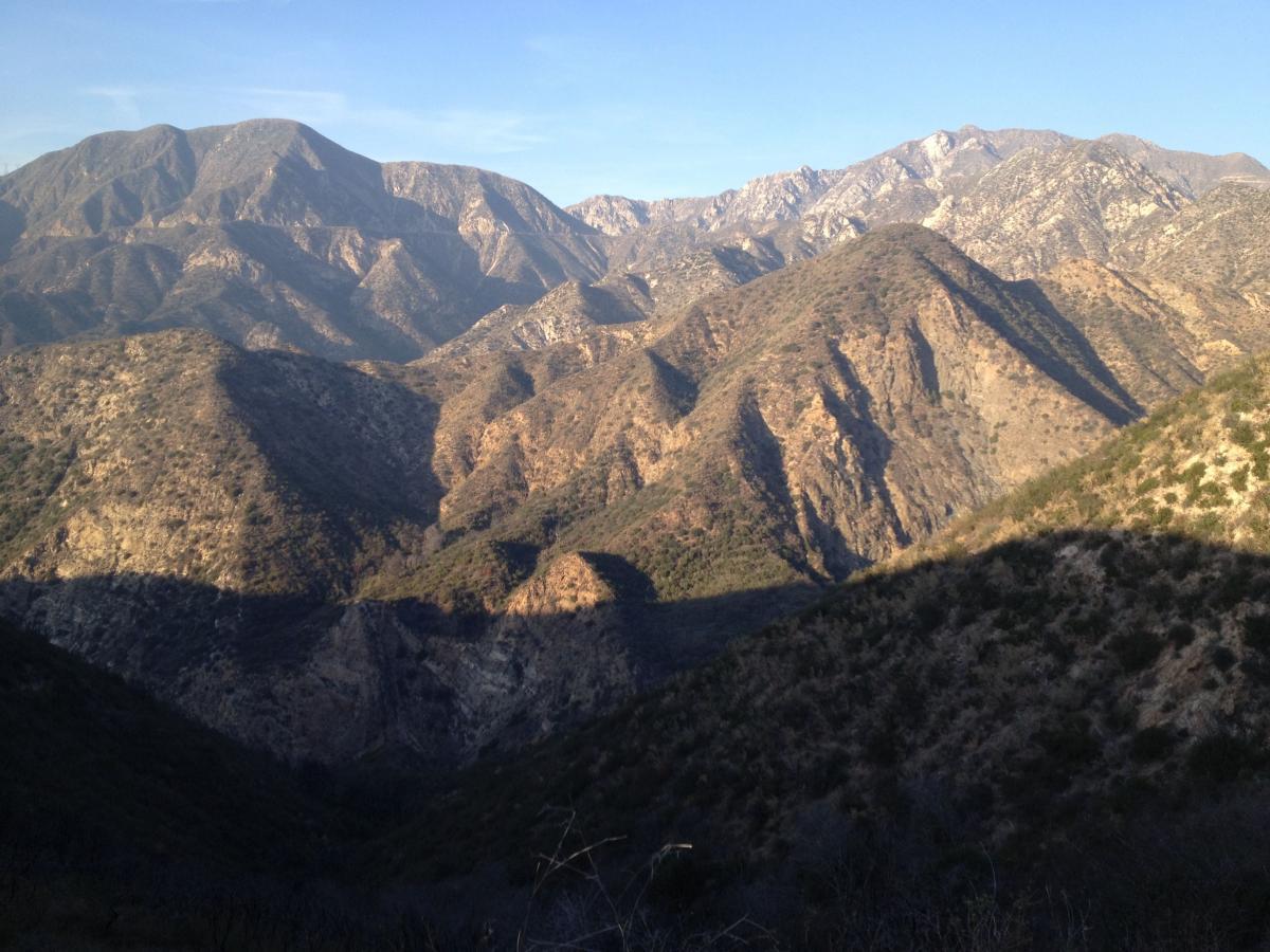 A panoramic view of a mountainous landscape featuring rugged peaks and rolling hills. The scene captures the natural beauty of the terrain, with varying shades of brown and green vegetation in the foreground and rocky summits in the background, under a clear blue sky. Shadows created by the mountains add depth to the image. El Prieto mountain bike trail.