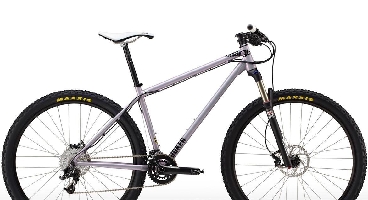 Charge Cooker 4: A mountain bike with a sleek silver frame, featuring a white seat, prominent Maxxis tires, and a front suspension fork. The bike is positioned against a plain white background, highlighting its design and components.