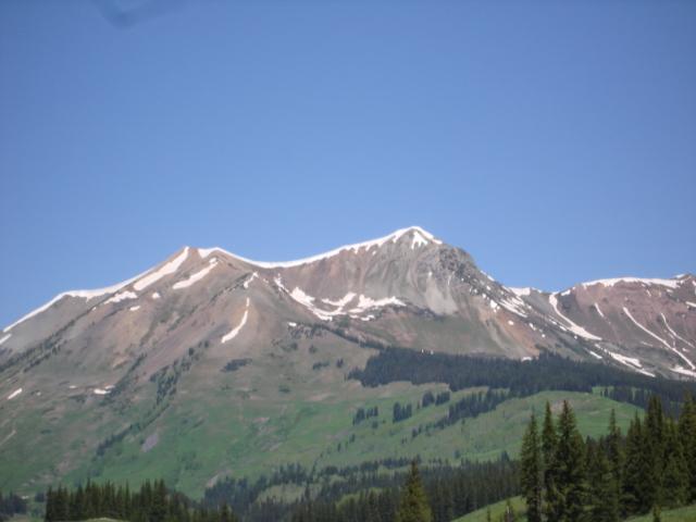 A rugged mountain landscape under a clear blue sky, featuring snow-capped peaks and lush green slopes. The foreground shows dense evergreen trees, while the backdrop highlights the rocky, multicolored mountain ridges. Trail 401 mountain bike trail.
