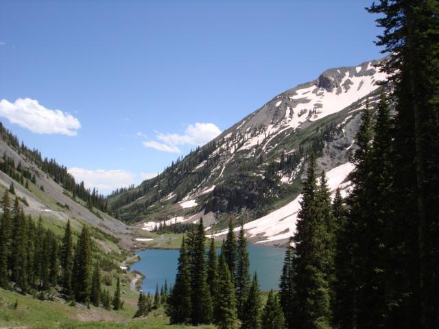A picturesque landscape featuring a serene lake surrounded by lush greenery and towering mountains. Snow-capped peaks rise in the background under a clear blue sky, with scattered clouds adding to the tranquil atmosphere. Evergreen trees frame the scene, enhancing the natural beauty of the area. Trail 401 mountain bike trail.