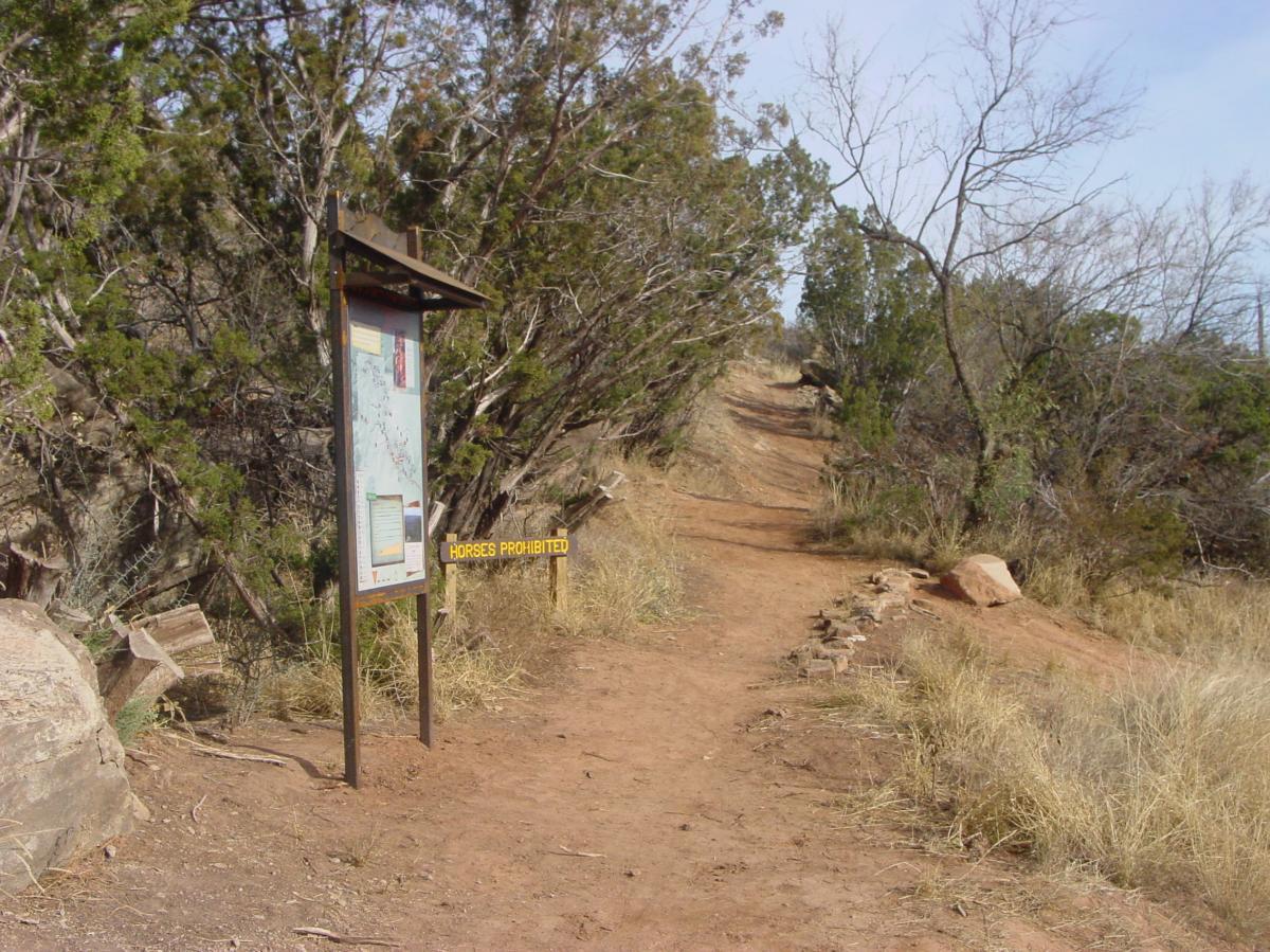 A dirt trail surrounded by bushes and trees, featuring a trail map sign and a yellow "Horses Prohibited" sign. The pathway leads into the distance, flanked by rocky terrain and dry grass. Palo Duro Canyon mountain bike trail.