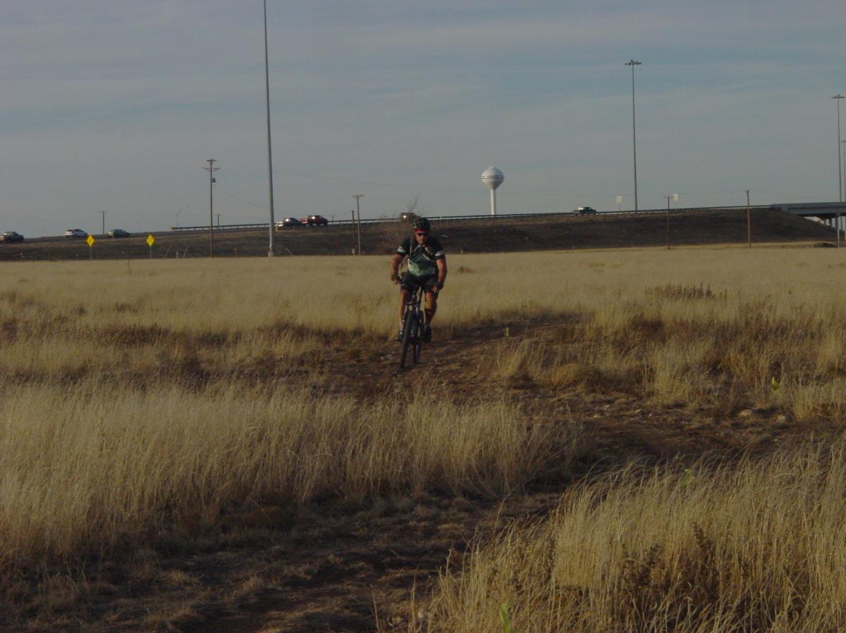 A person riding a mountain bike along a dirt path in a grassy field, with a highway and water tower visible in the background under a cloudy sky. Buffalo Trails mountain bike trail.