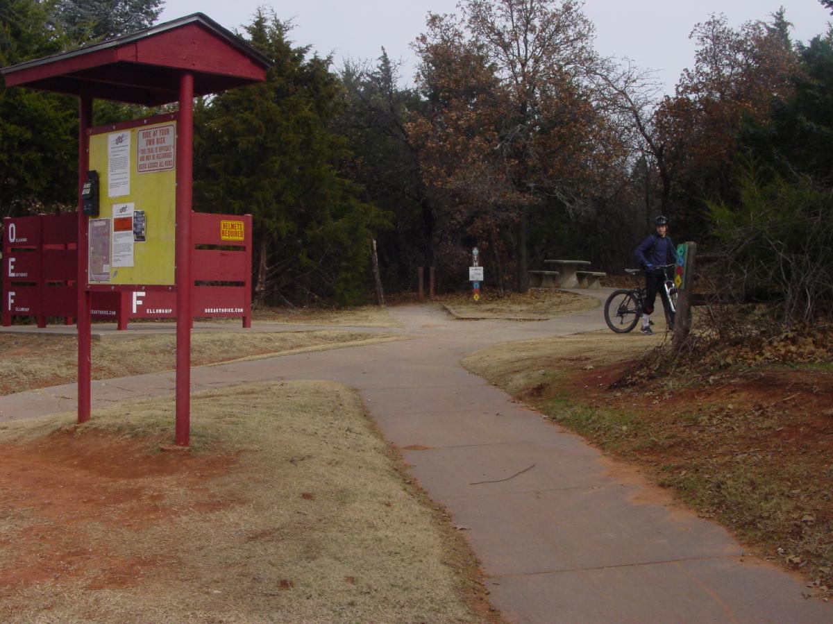 A bike trail intersection featuring a red information kiosk displaying trail maps and rules, with a cyclist in a helmet standing next to a mountain bike. The scene is surrounded by trees and a grassy area, under a cloudy sky. Lake Stanley Draper mountain bike trail.
